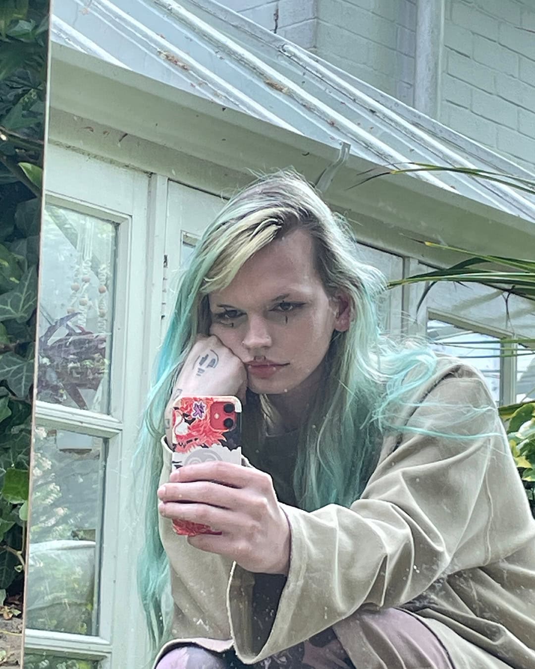Adult female poses with a cell phone in front of a reflective surface in an outdoor greenhouse setting with natural light.