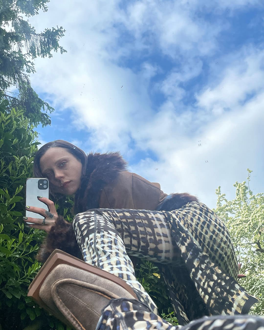 Woman in patterned pants and brown jacket takes selfie in outdoor setting with greenery and sky visible in background, low-angle perspective.