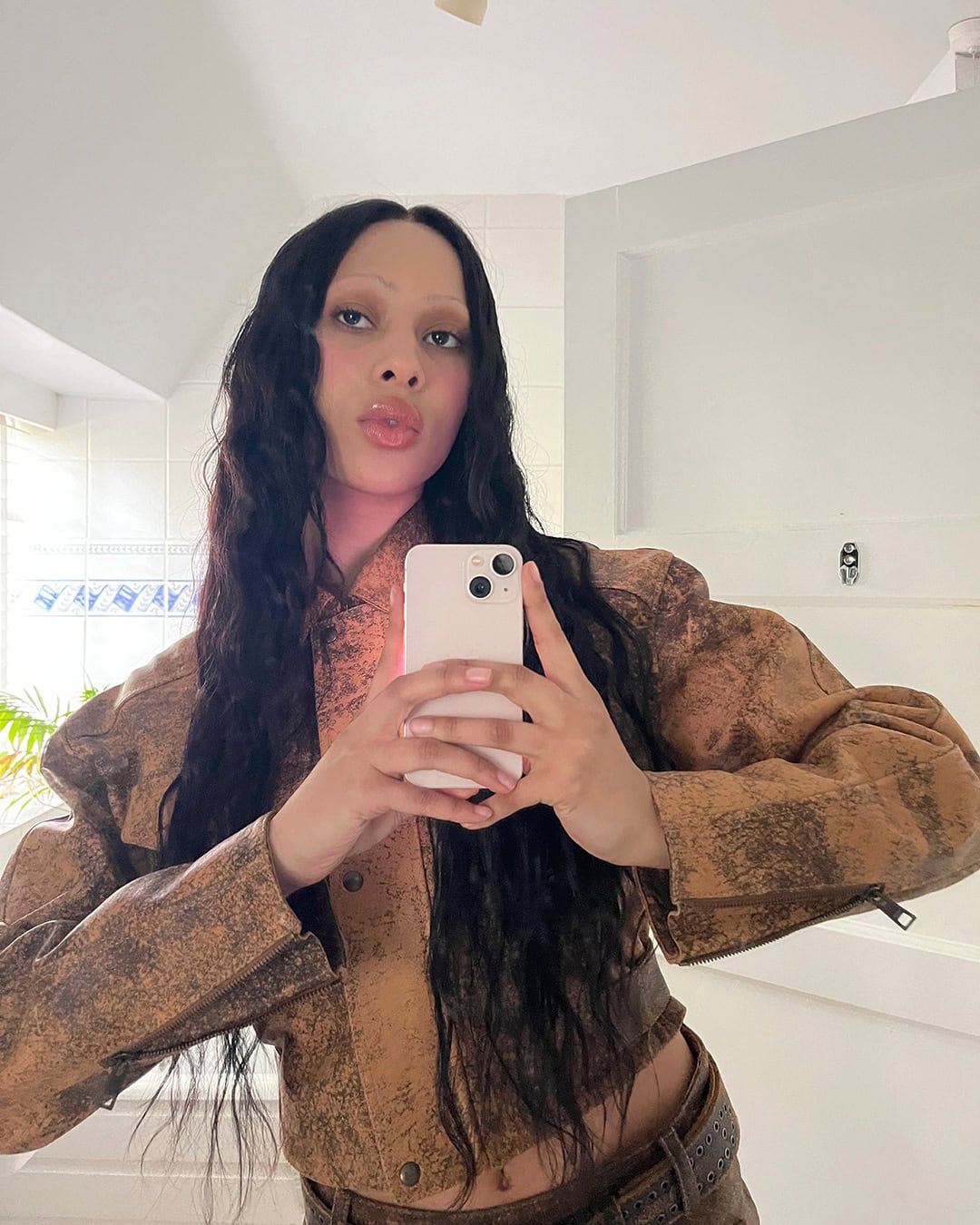 Young woman takes a selfie in a tan jacket and dark hair. She holds her phone and poses in an indoor setting with white walls.