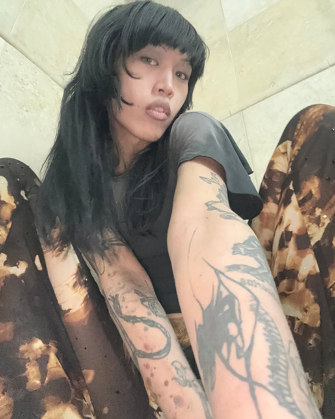 A woman with black hair and bangs looks at the camera with neutral expression in an indoor setting. Tattoos are visible on her arm.