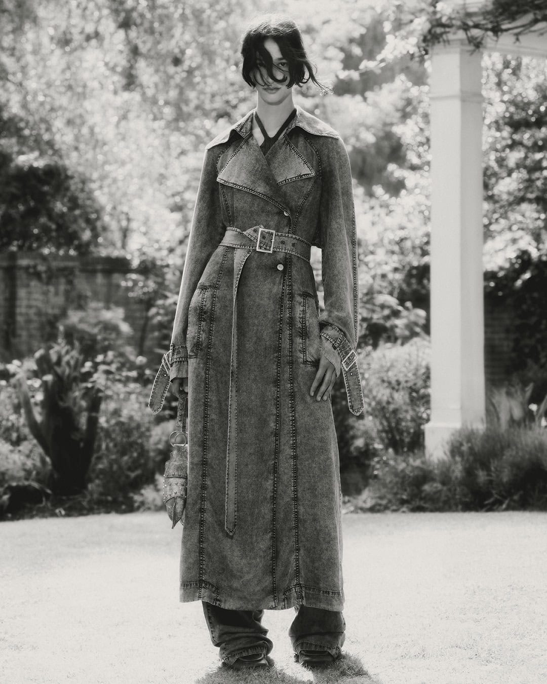 Monochrome shot of a woman in a long denim trench coat standing in a garden with a white pergola and greenery, capturing a fashion editorial style.