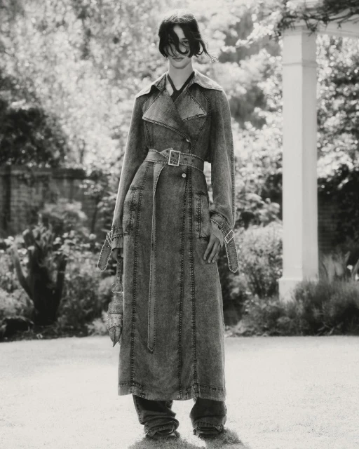 Monochrome shot of a woman in a long denim trench coat standing in a garden with a white pergola and greenery, capturing a fashion editorial style.