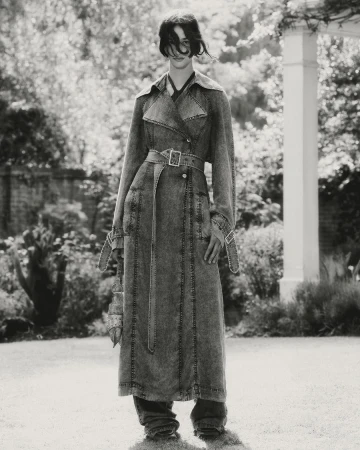 Monochrome shot of a woman in a long denim trench coat standing in a garden with a white pergola and greenery, capturing a fashion editorial style.