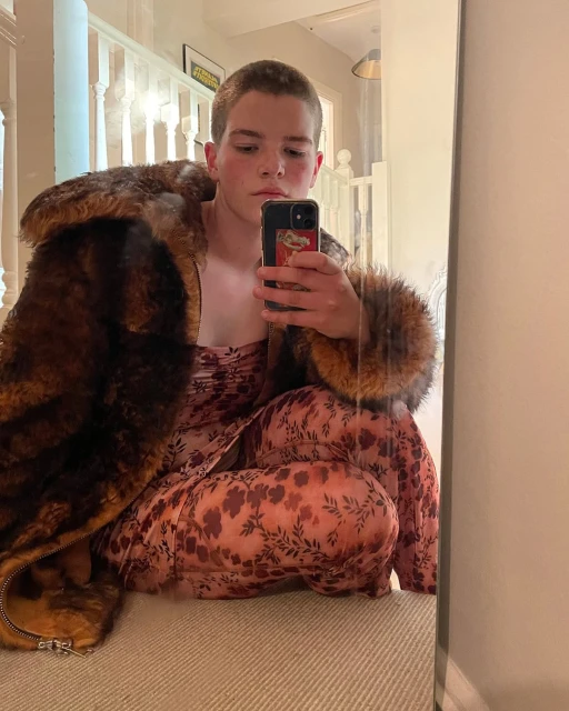 Person wearing a floral dress and fur coat takes a mirror selfie inside a hallway with neutral-toned walls and beige carpet.