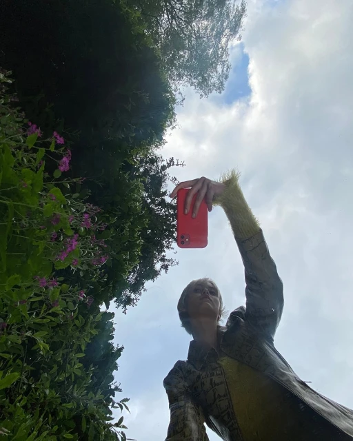 A woman in patterned jacket takes a photo with a red phone outdoors against a backdrop of trees and sky on a sunny day.