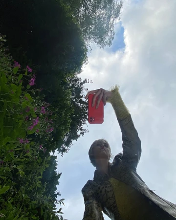 A woman in patterned jacket takes a photo with a red phone outdoors against a backdrop of trees and sky on a sunny day.