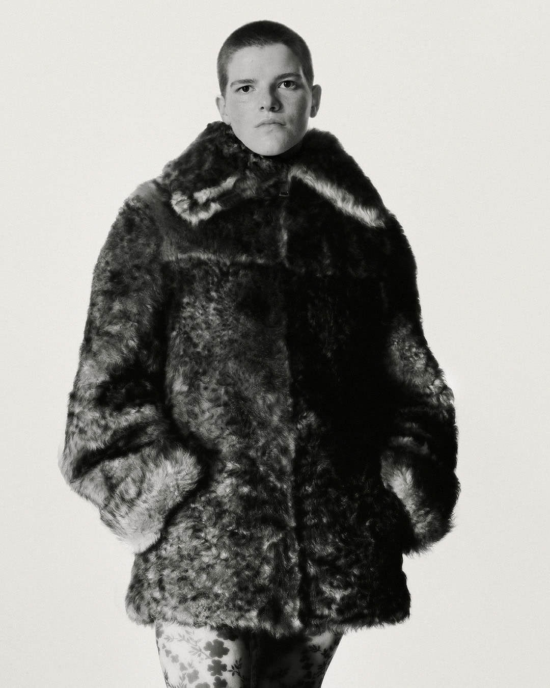Model with a shaved head wearing a dark textured faux fur coat, standing against a plain white background in a studio setting.