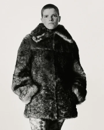Model with a shaved head wearing a dark textured faux fur coat, standing against a plain white background in a studio setting.