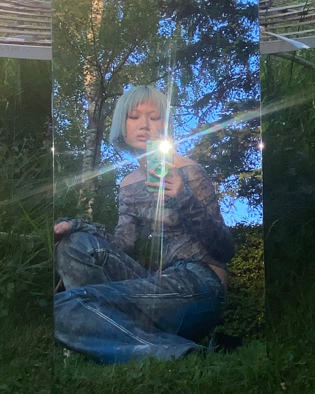 Young woman with pastel hair poses outdoors near tall mirror reflecting trees and sky, wearing a dark lace top and light wash jeans.