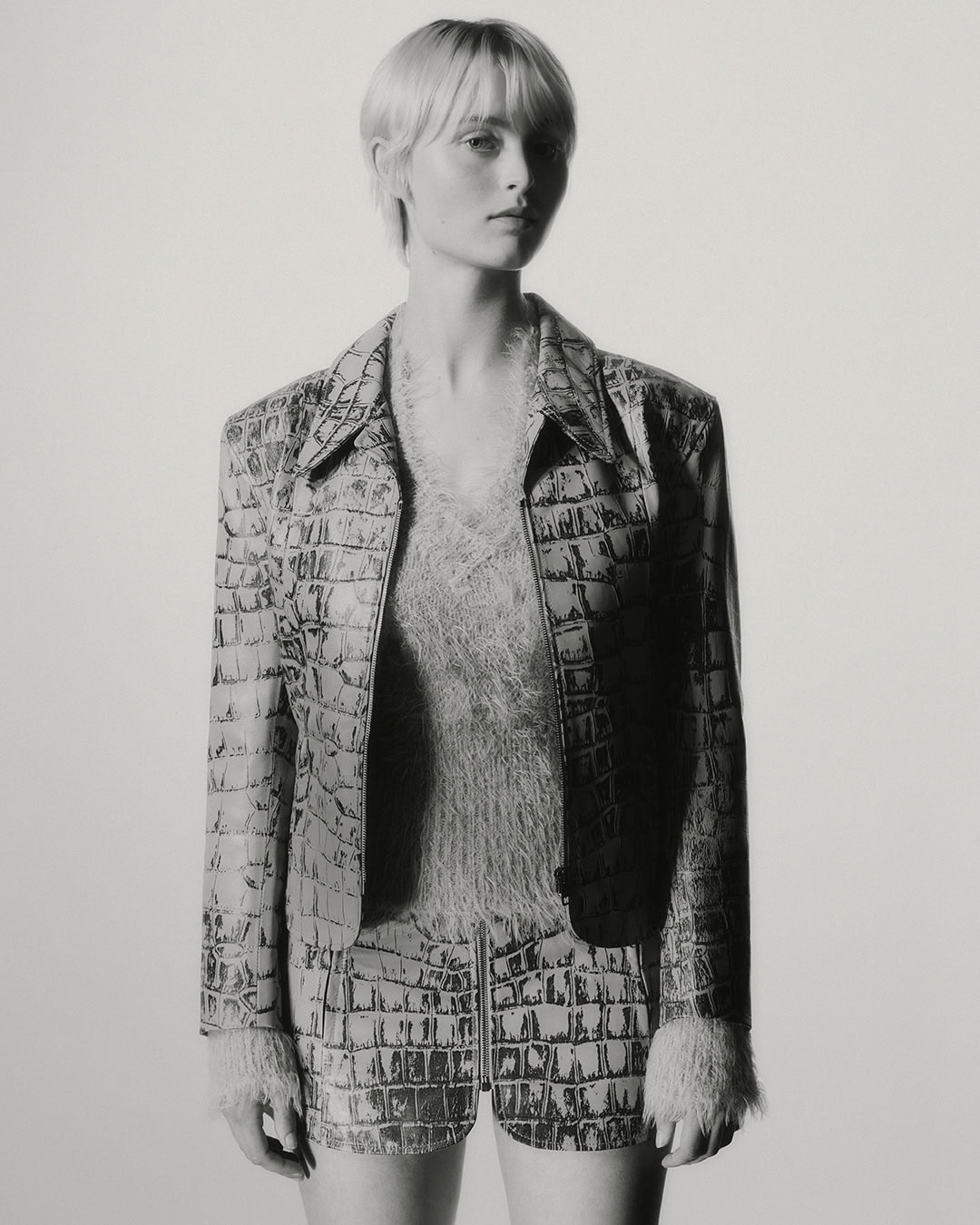 Portrait of a slender adult female model with fair skin, wearing a reptile-pattern jacket and shorts in a studio setting.
