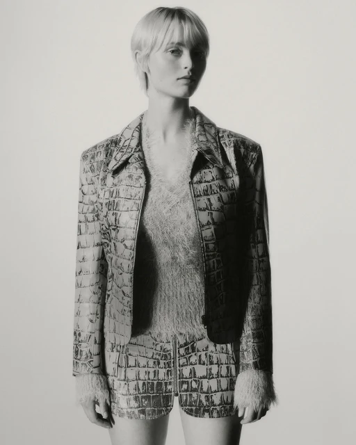 Portrait of a slender adult female model with fair skin, wearing a reptile-pattern jacket and shorts in a studio setting.