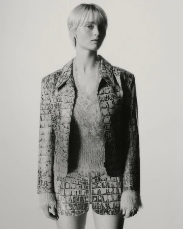 Portrait of a slender adult female model with fair skin, wearing a reptile-pattern jacket and shorts in a studio setting.