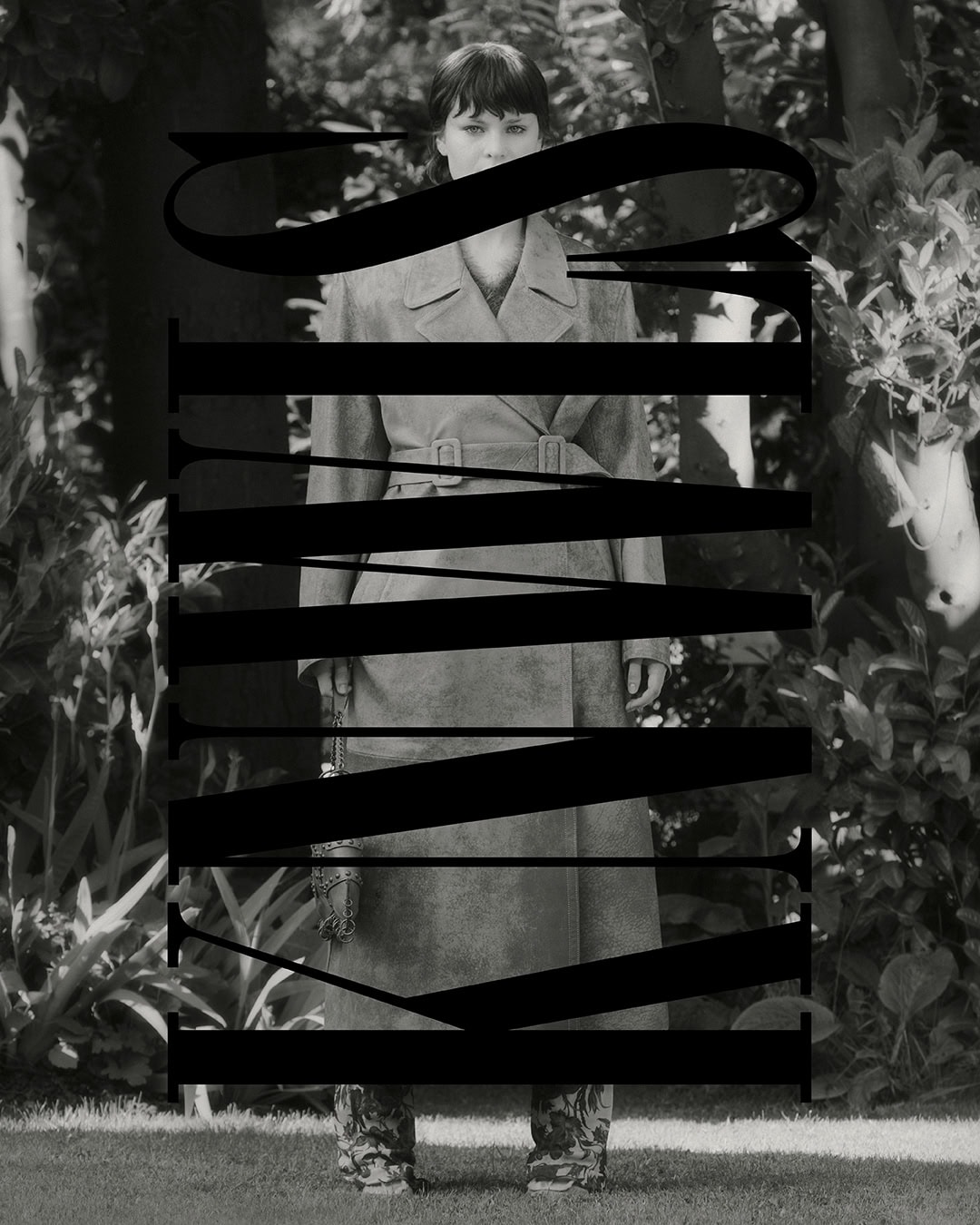 Monochrome portrait of a woman in a trench coat standing outdoors amongst lush foliage, partially obscured by large black typography.