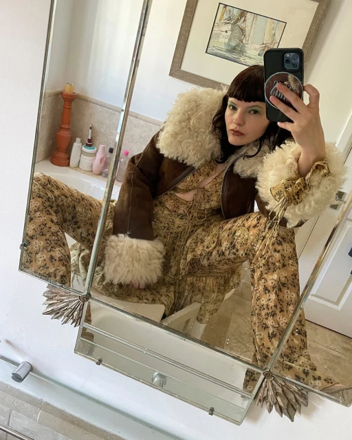 Woman with dark hair wearing a floral print dress and shearling coat in an indoor bathroom setting, reflected in a mirror.