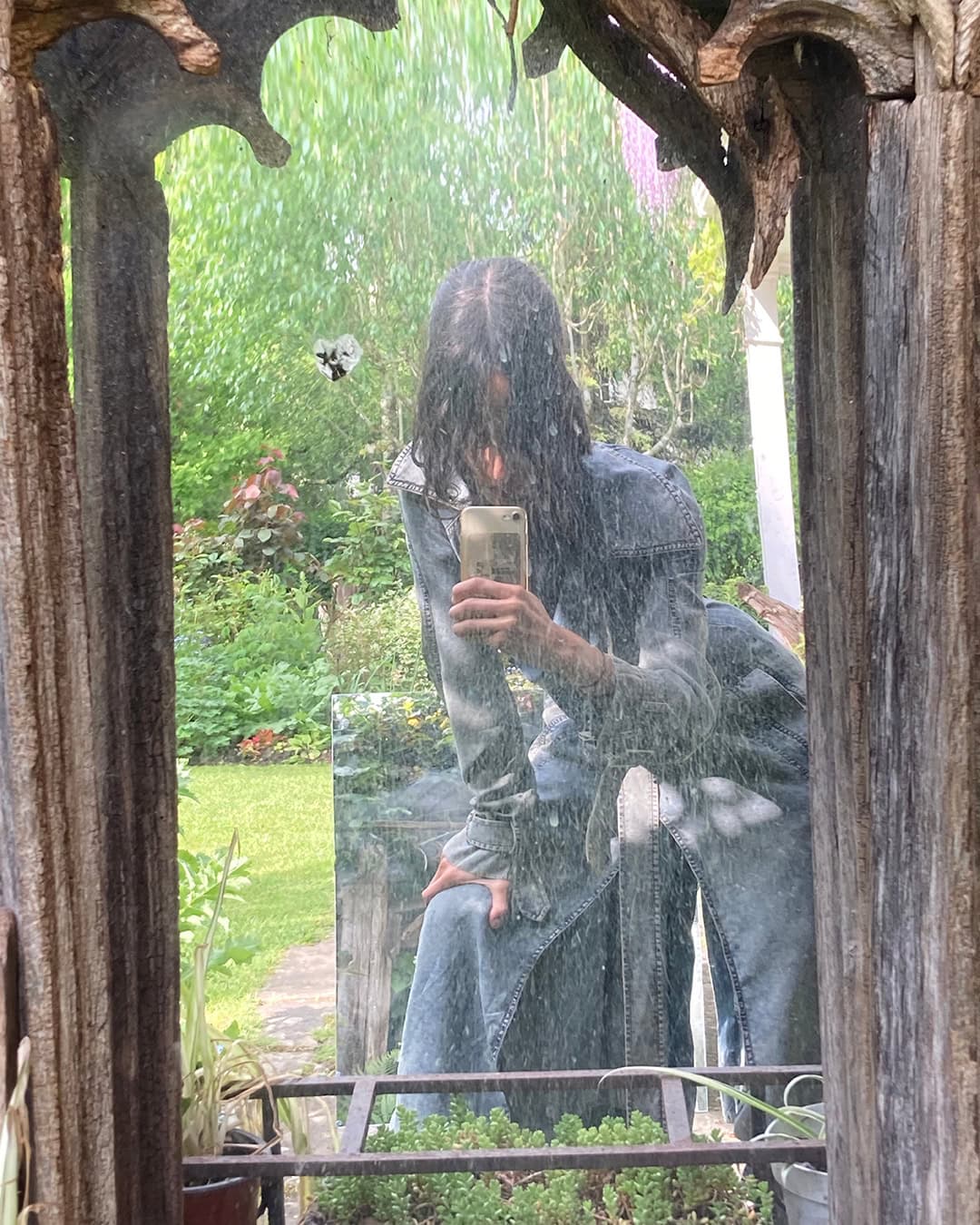 Adult poses for a selfie in a rustic wood framed mirror that reflects a verdant outdoor garden setting on a rainy day.