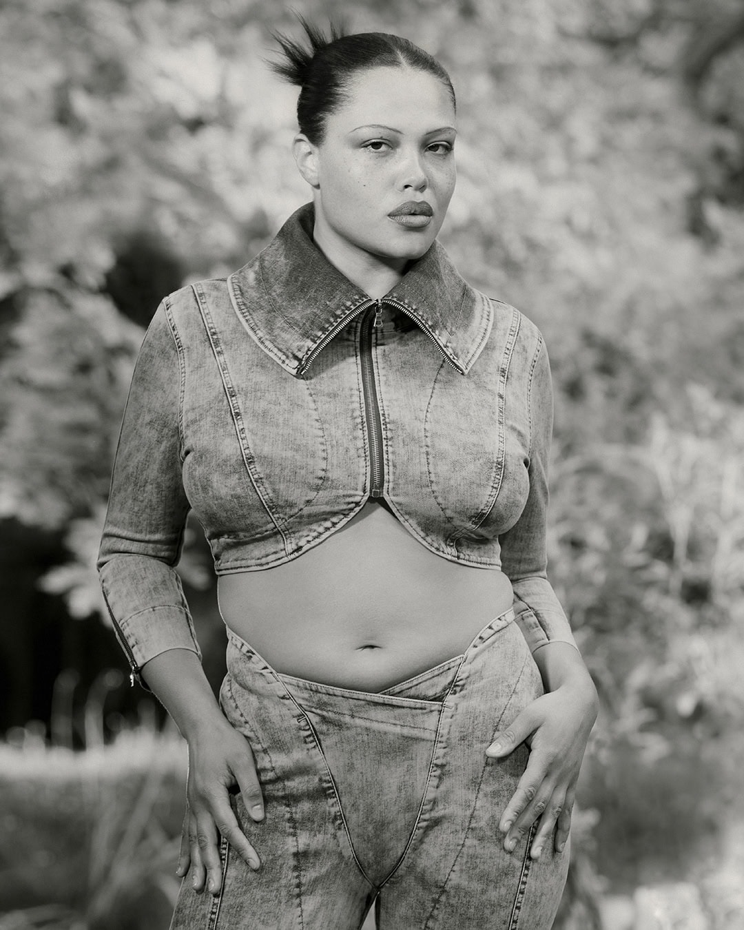 Model Alva Claire posing in a cropped denim jacket and matching jeans against blurred foliage for KNWLS.