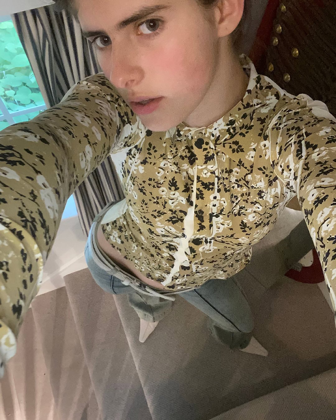 Portrait of a fair-skinned woman in a floral top, shot from a high angle in an indoor environment with natural light from a nearby window.