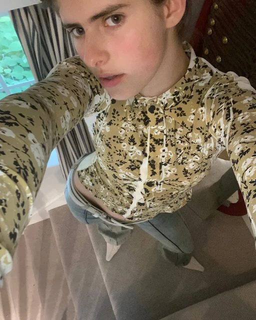 Portrait of a fair-skinned woman in a floral top, shot from a high angle in an indoor environment with natural light from a nearby window.