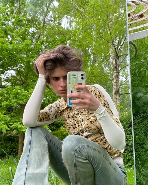 Teenager poses with a mobile phone, seated outdoors against a lush green backdrop, wearing casual clothing in a reflective mirror scene.