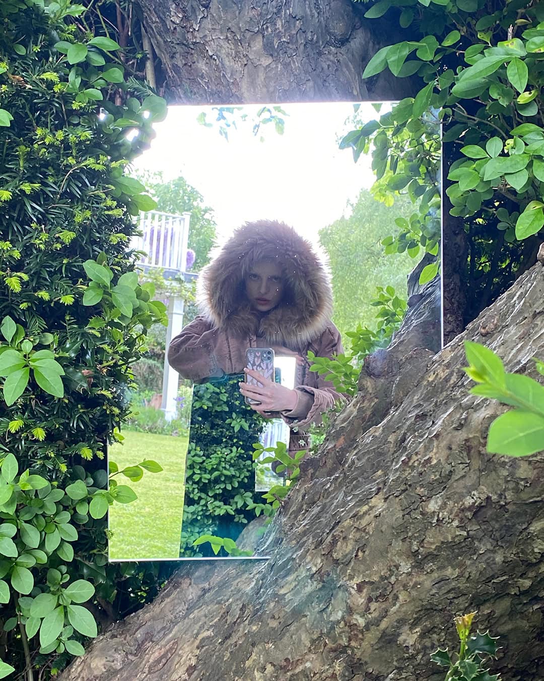 Person takes selfie in a mirror framed by a tree trunk and green leaves, wearing a fur-trimmed hooded jacket outdoors.