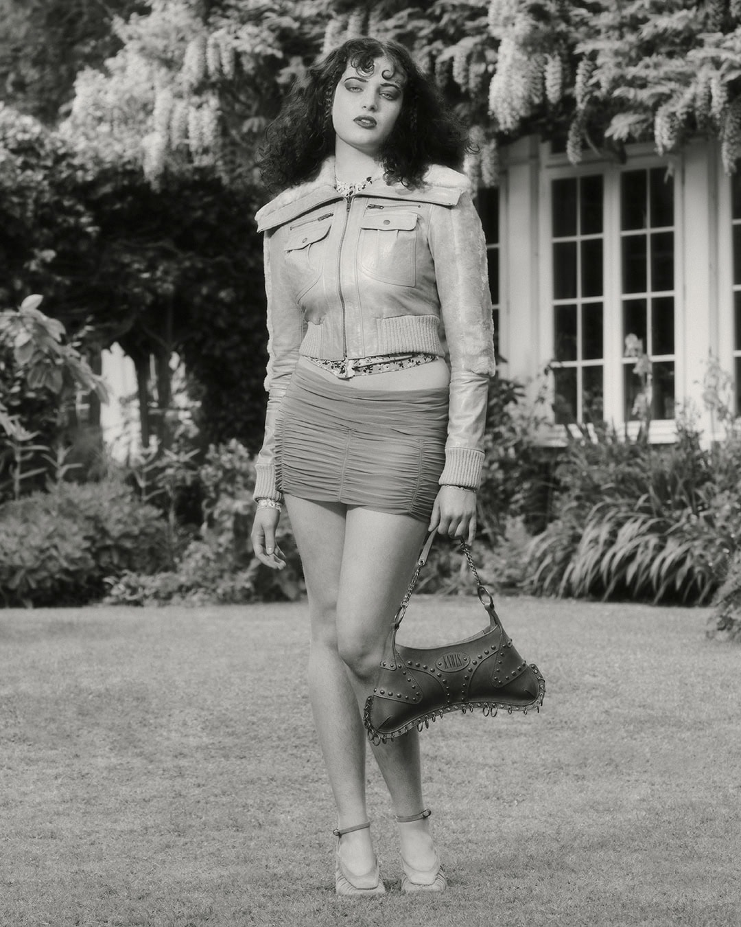 Full-length shot of a woman in a jacket and mini-skirt posing with a handbag in a yard with a house and foliage background in black and white.