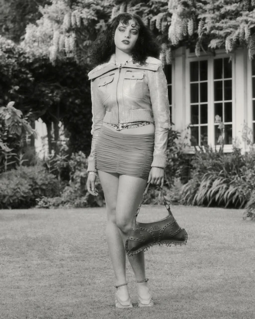 Full-length shot of a woman in a jacket and mini-skirt posing with a handbag in a yard with a house and foliage background in black and white.