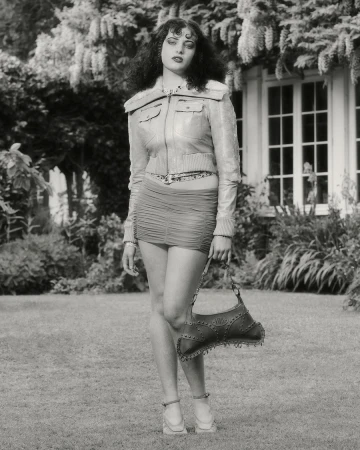 Full-length shot of a woman in a jacket and mini-skirt posing with a handbag in a yard with a house and foliage background in black and white.