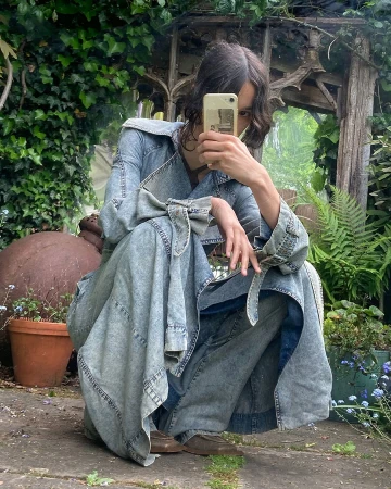 A person in a denim outfit crouches in a lush garden setting, capturing a photo with a smartphone, surrounded by greenery and plants.