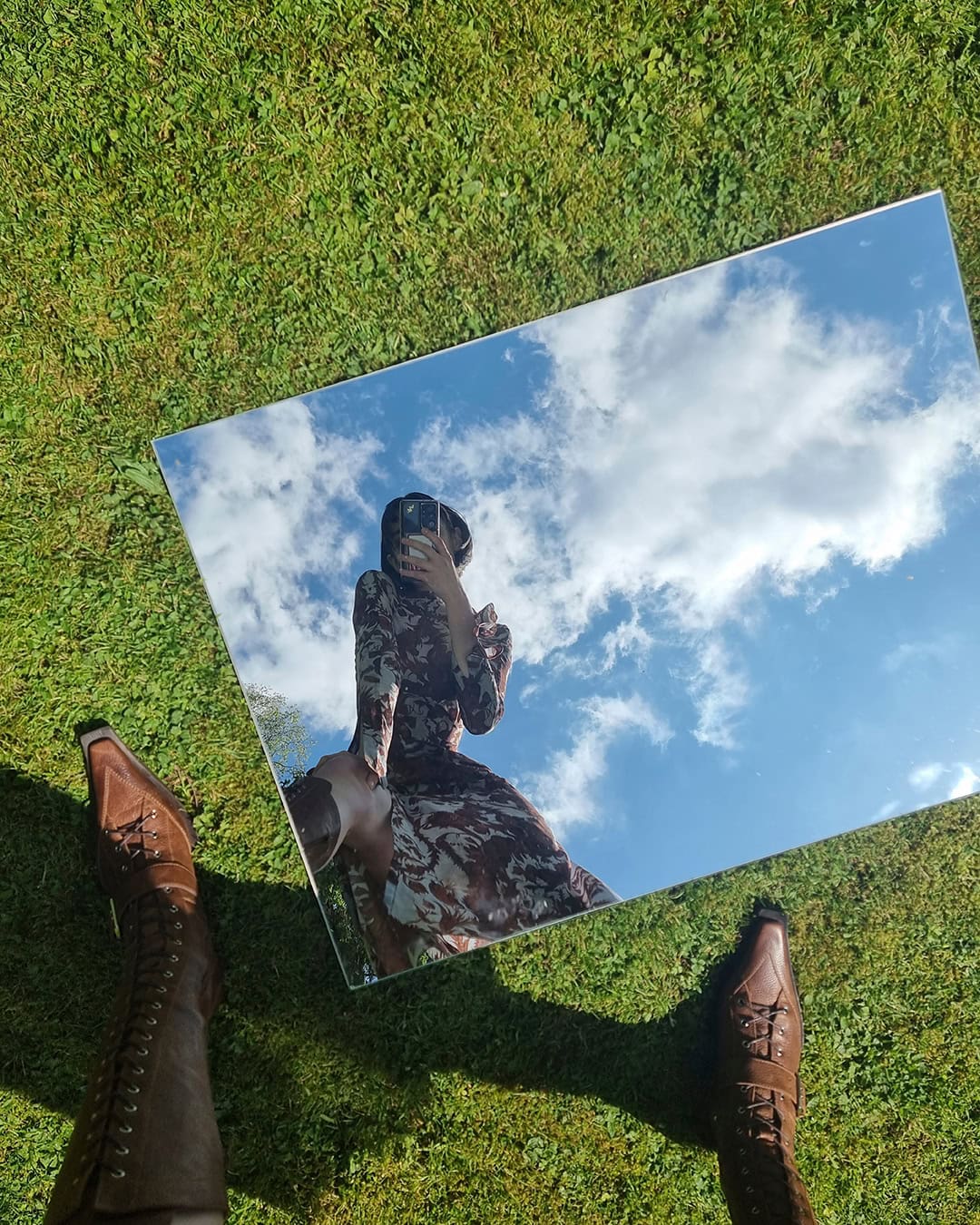 Top-down view of brown lace-up boots standing on green grass beside a mirror reflecting a person in a printed dress and blue sky.