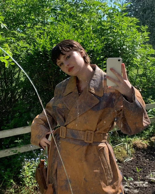 A young woman with short brunette hair takes a selfie outside, wearing a brown trench coat in front of lush greenery and bright sun.