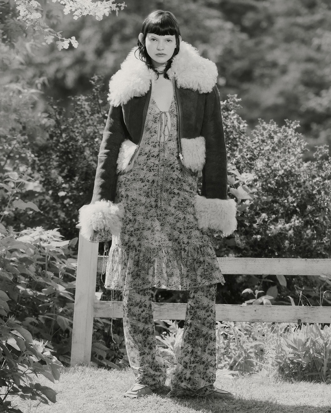 Monochrome portrait of a woman in a floral dress and shearling jacket outdoors. She is standing in a garden with a fence behind her.