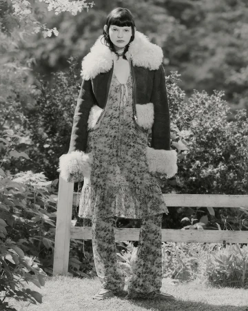 Monochrome portrait of a woman in a floral dress and shearling jacket outdoors. She is standing in a garden with a fence behind her.