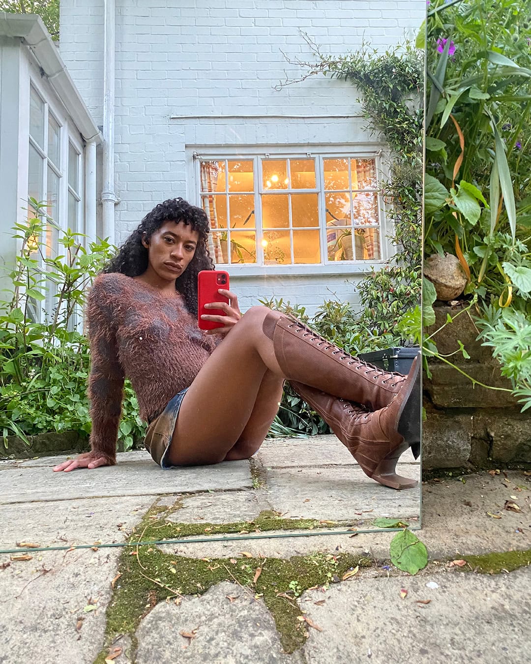 Adult female sits outside by a building and mirror, wearing a brown sweater, shorts, and boots, holding a red cell phone.