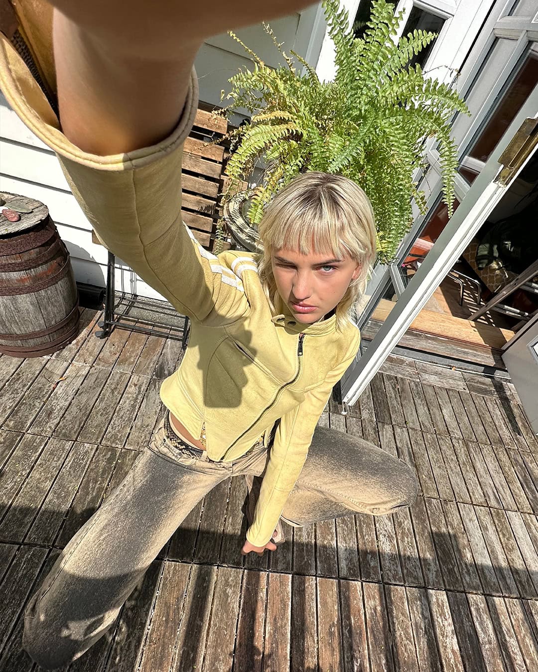 Blonde adult poses, squatting on a wood deck in the sun, wearing a fitted yellow jacket and acid-wash jeans; barrel and plant behind.