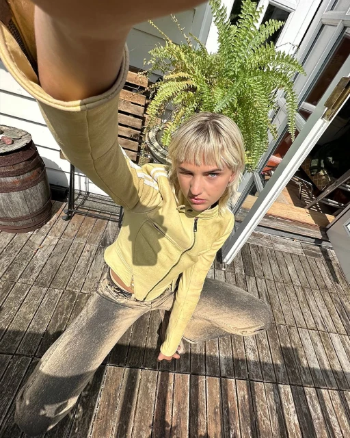 Blonde adult poses, squatting on a wood deck in the sun, wearing a fitted yellow jacket and acid-wash jeans; barrel and plant behind.