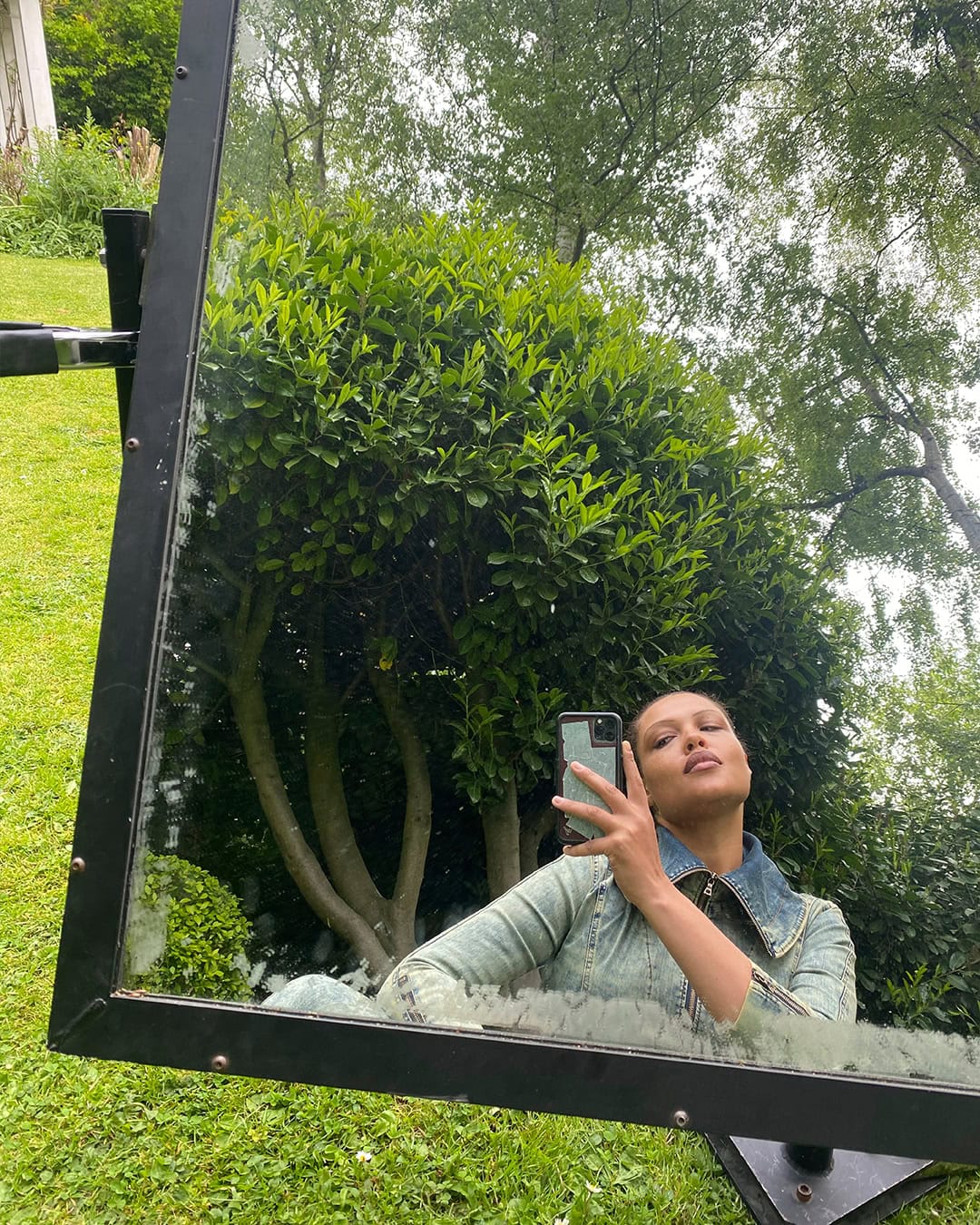 A person takes a selfie reflected in a large mirror outdoors surrounded by green grass and foliage, wearing a denim jacket.