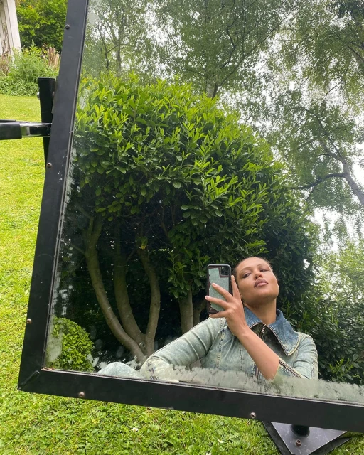 A person takes a selfie reflected in a large mirror outdoors surrounded by green grass and foliage, wearing a denim jacket.