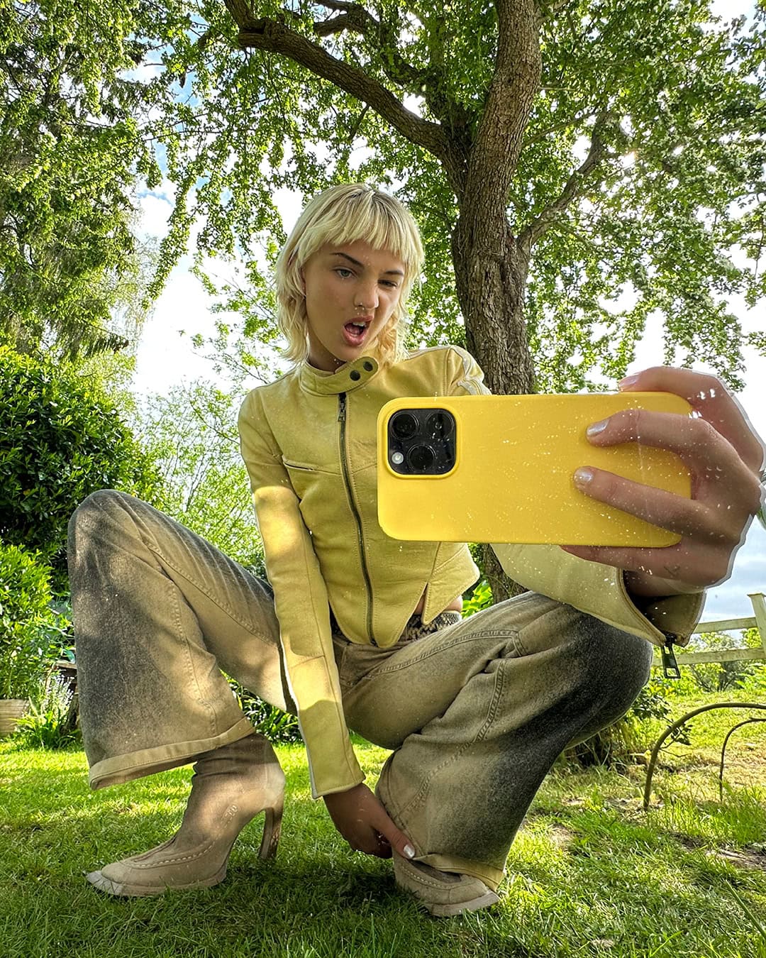 A young adult woman poses for a selfie outdoors in a yellow jacket, patterned pants, and heeled boots, surrounded by green foliage.