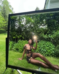 Model reclining on a green lawn taking a mirror reflection photo wearing a printed two-piece outfit and brown knee-high lace-up boots.