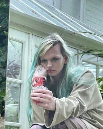 Person with long pastel green hair taking a mirror selfie on a smartphone wearing a beige oversized top near a greenhouse window.