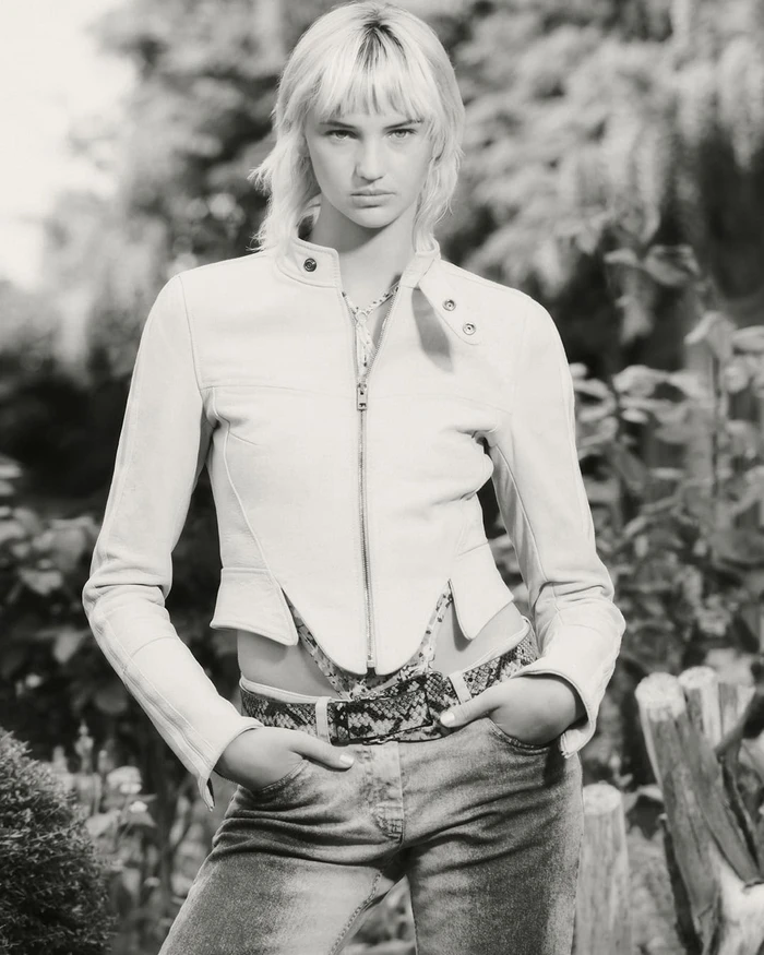 Model wearing a cropped white leather jacket and patterned belt outdoors in a black and white portrait.