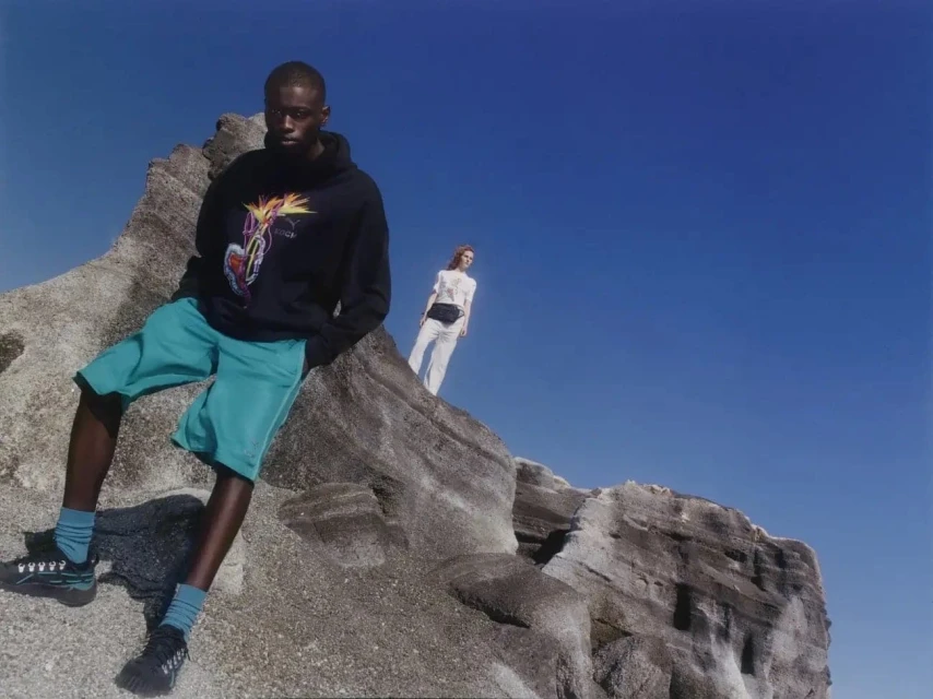 A person in a hoodie and shorts leans on a rock formation against a blue sky, with another person standing in the background.