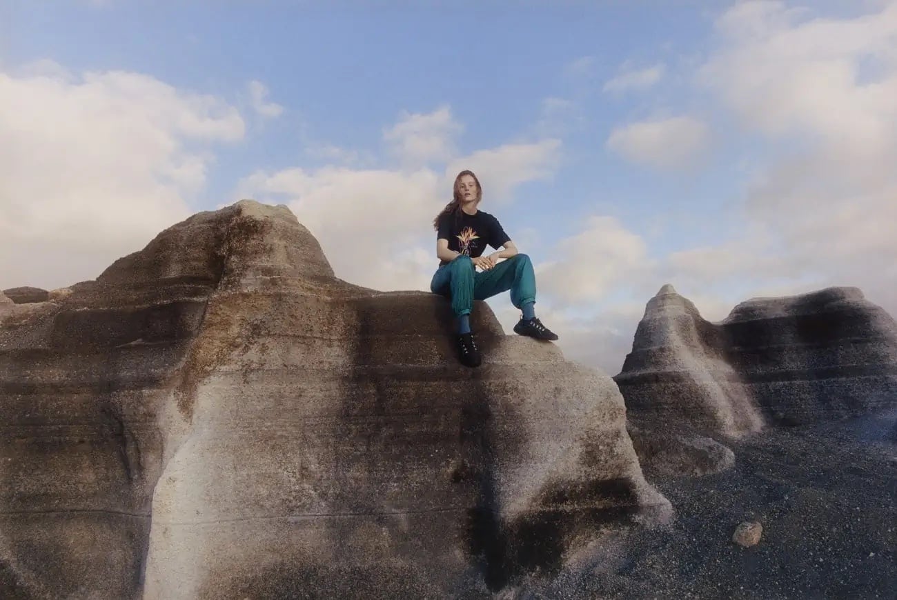Adult female seated on rock formation under blue sky with clouds, wearing a black t-shirt and blue-green pants in an outdoor environment.