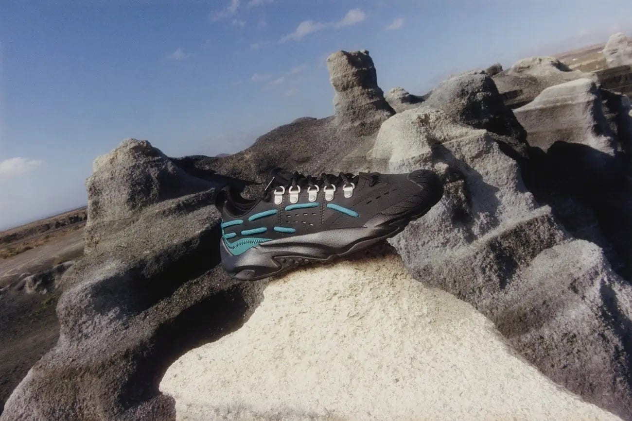 Elevated view of a black athletic shoe with teal accents on a textured rock formation against a partly cloudy blue sky backdrop.