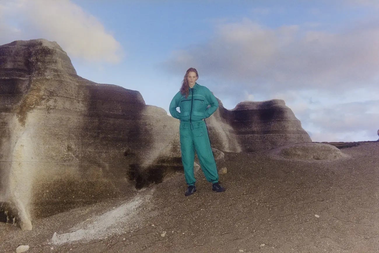 A woman in a teal jumpsuit stands on a sandy dune with her hands on her hips, with a large, weathered rock formation behind her.