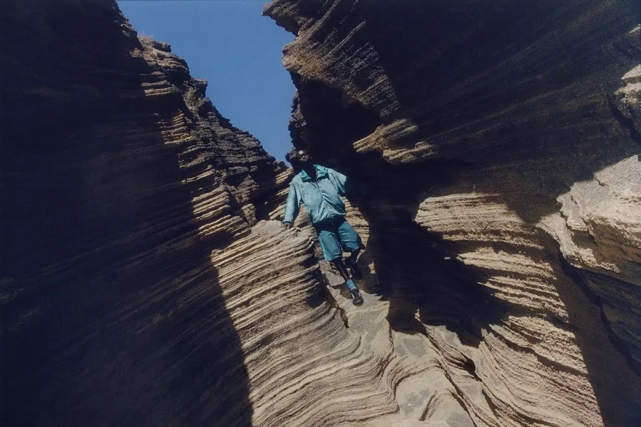 Man in a blue jacket climbs layered rock formations with a blue sky visible above. Outdoor adventure and natural rock features.