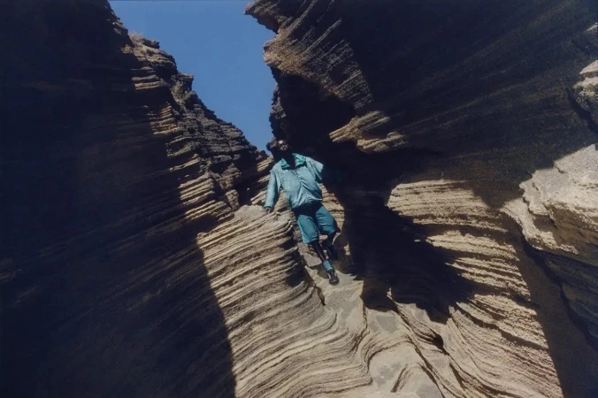 Man in a blue jacket climbs layered rock formations with a blue sky visible above. Outdoor adventure and natural rock features.
