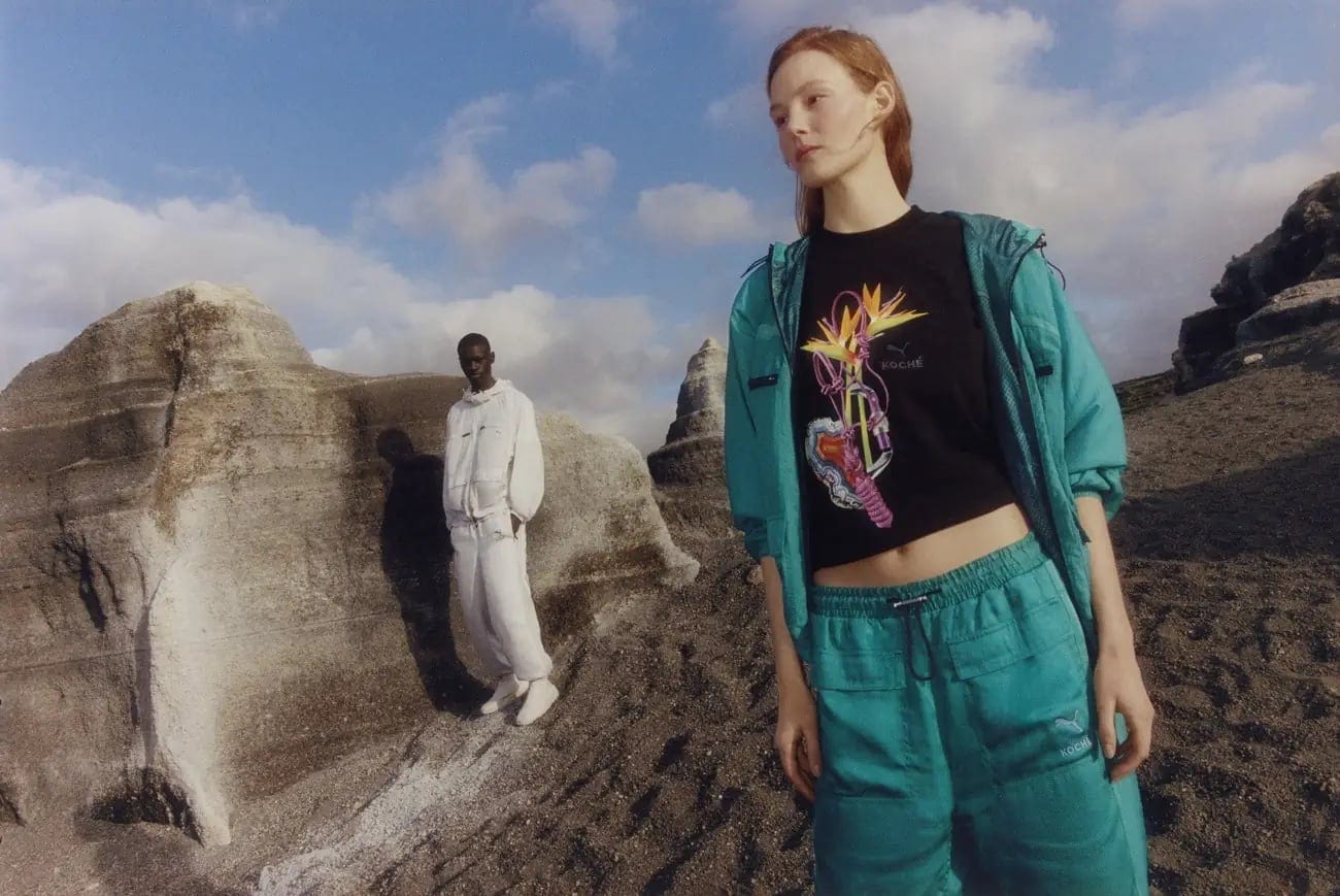 Two fashion models pose outdoors in sportswear on a rocky landscape under a blue, cloudy sky. One wears white, the other wears teal.