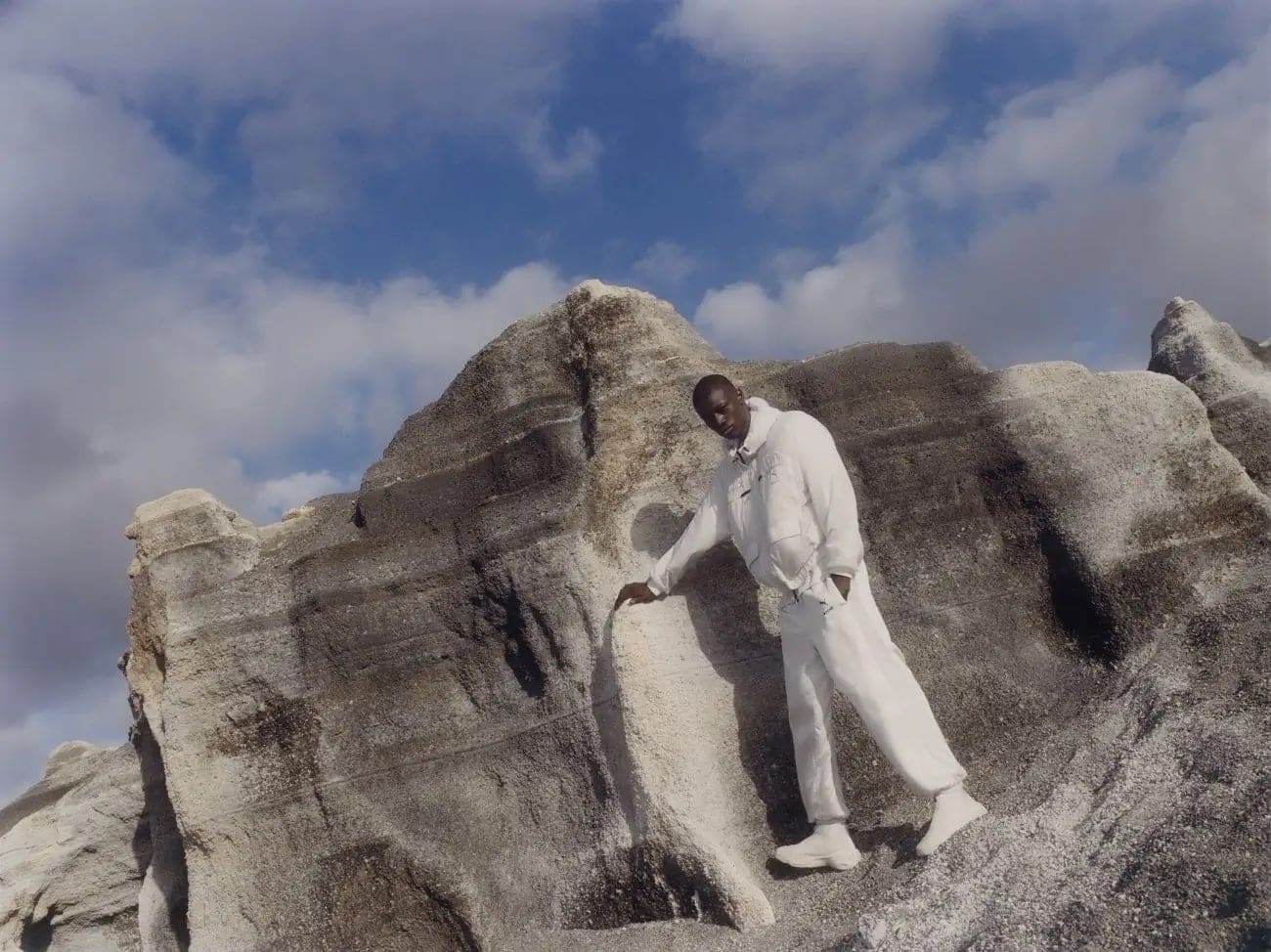 An adult male model poses in a white outfit on a textured rock formation under a cloudy sky, fashion photography.