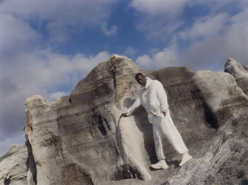 An adult male model poses in a white outfit on a textured rock formation under a cloudy sky, fashion photography.